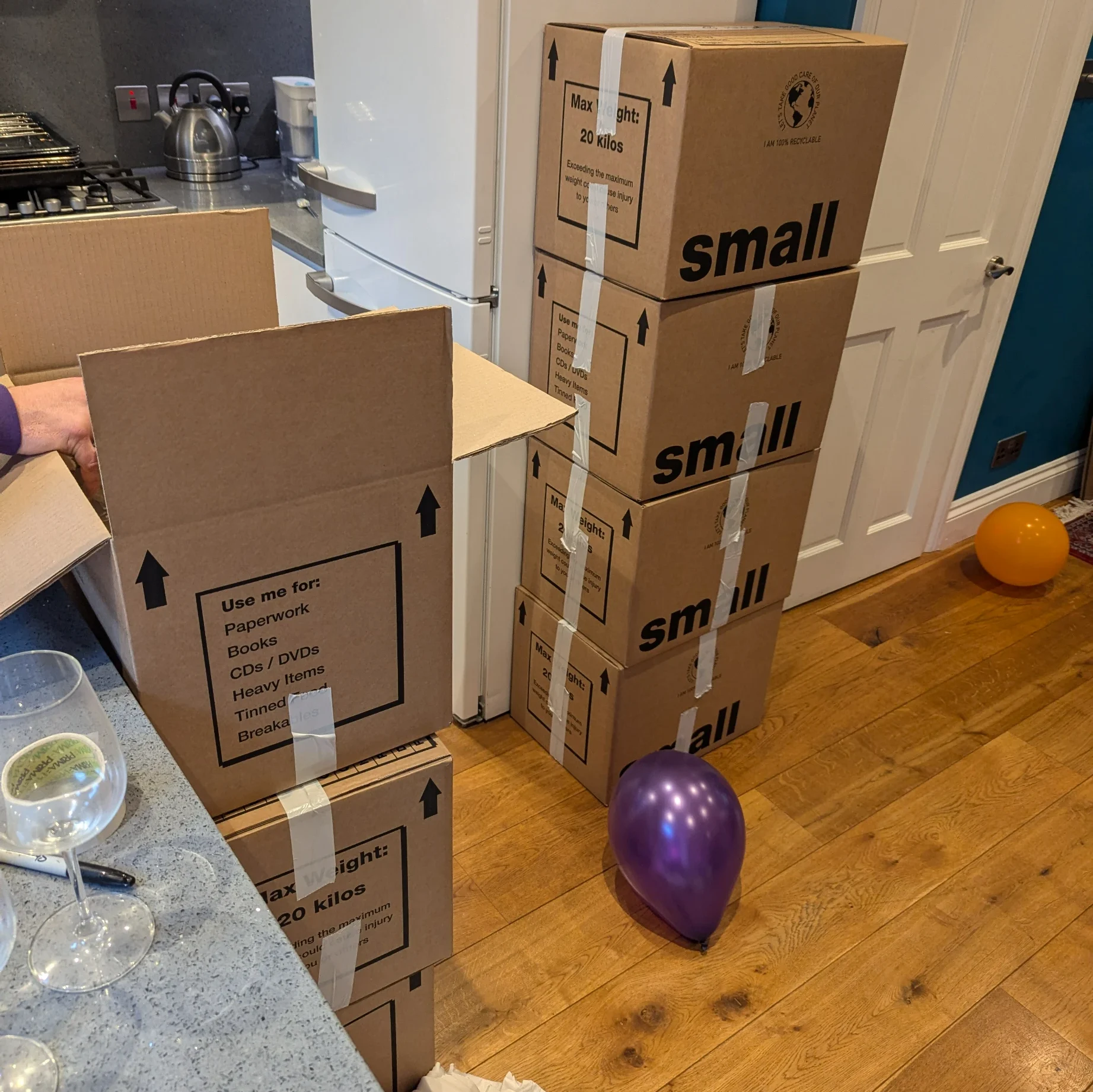 Moving boxes stacked in a London home, with a purple balloon on the floor and wine glasses on the kitchen counter